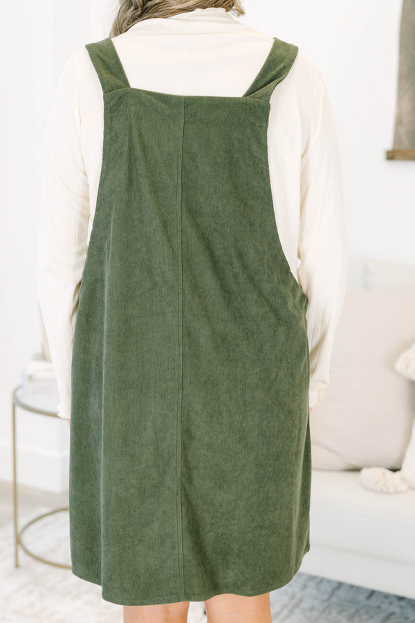 Plus size olive green corduroy overall dress with tie straps and front pockets over a cream top.