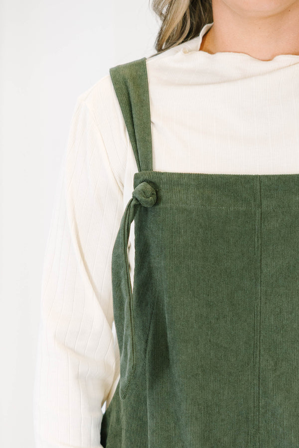 Plus size olive green corduroy overall dress with tie straps and front pockets over a cream top.