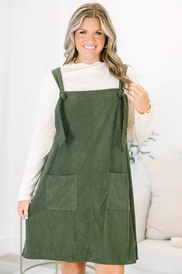 Plus size olive green corduroy overall dress with tie straps and front pockets over a cream top.