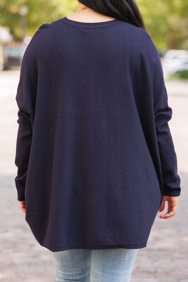 A woman smiles wearing a long-sleeved navy blue tunic with front pockets and light-wash jeans.