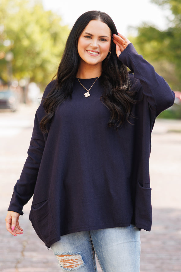 A woman smiles wearing a long-sleeved navy blue tunic with front pockets and light-wash jeans.