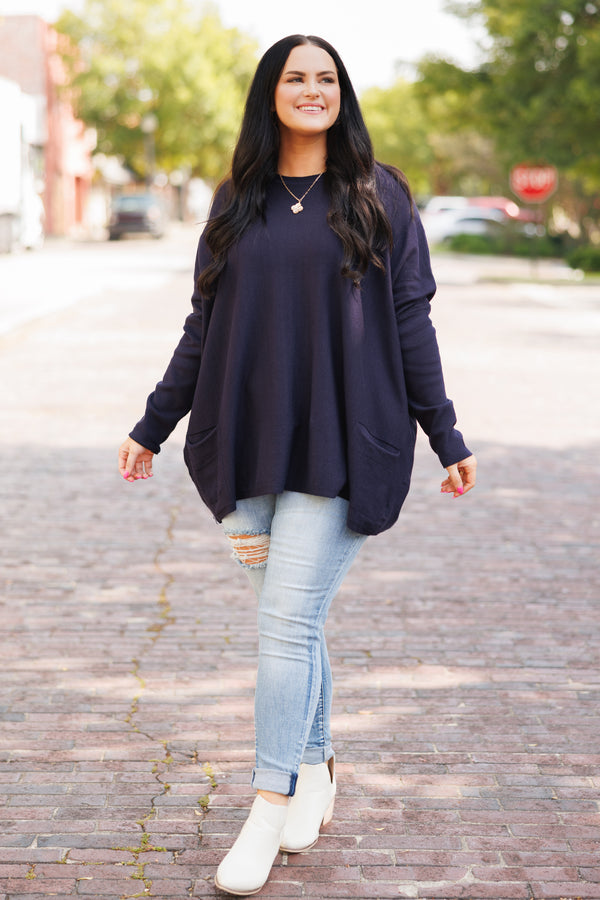 A woman smiles wearing a long-sleeved navy blue tunic with front pockets and light-wash jeans.