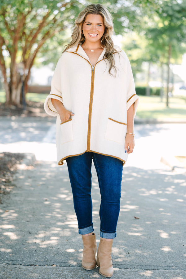 Plus size cream zip-up poncho sweater with brown trim, front pockets, and a flowy, relaxed fit.