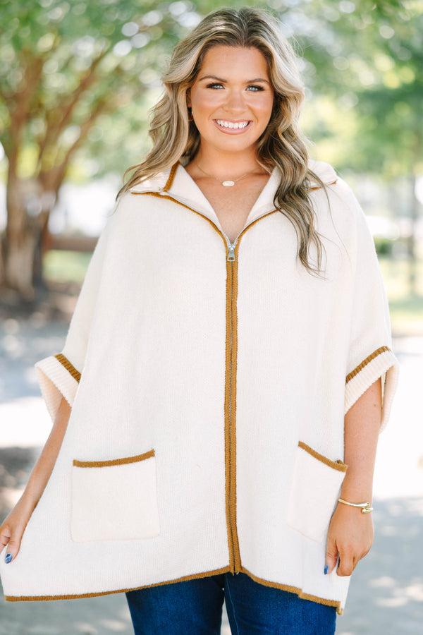 Plus size cream zip-up poncho sweater with brown trim, front pockets, and a flowy, relaxed fit.