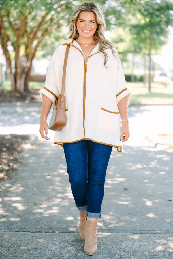 Plus size cream zip-up poncho sweater with brown trim, front pockets, and a flowy, relaxed fit.