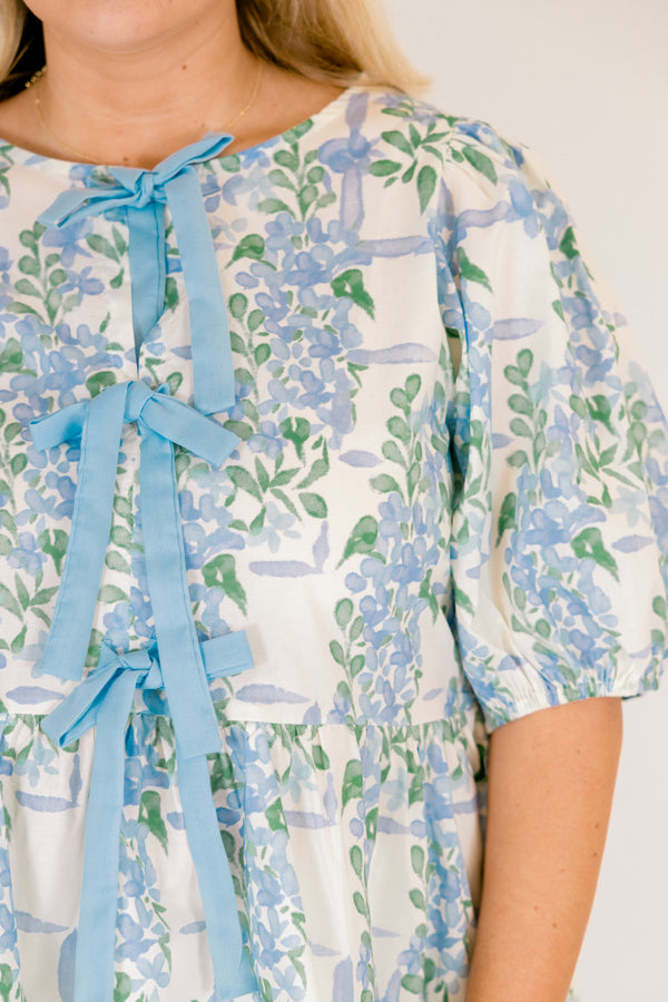 Chic Soul plus size clothing, white puff short sleeve babydoll top with light blue and green floral pattern and three blue bow ties down the middle of the top and cinching at the end of the sleeves